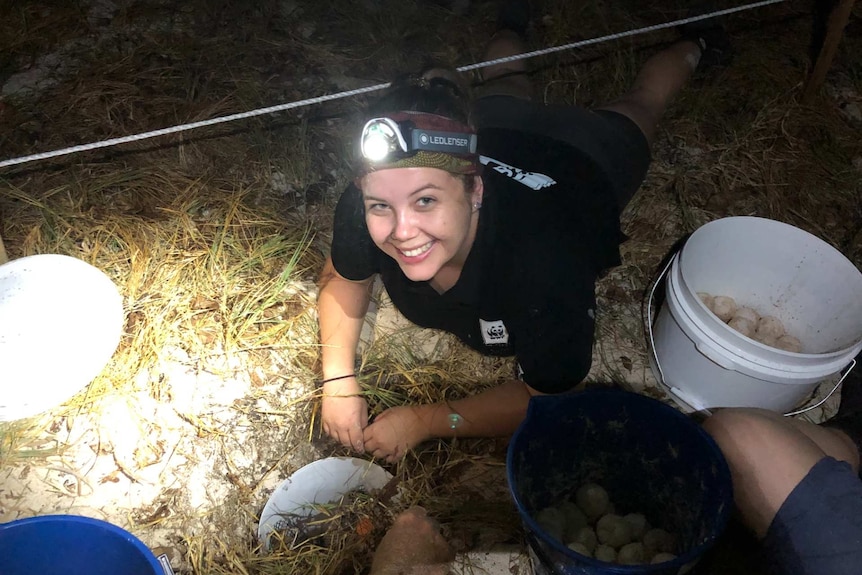 A lady wearing a head torch and lying on the sand with a bucket of turtle eggs.