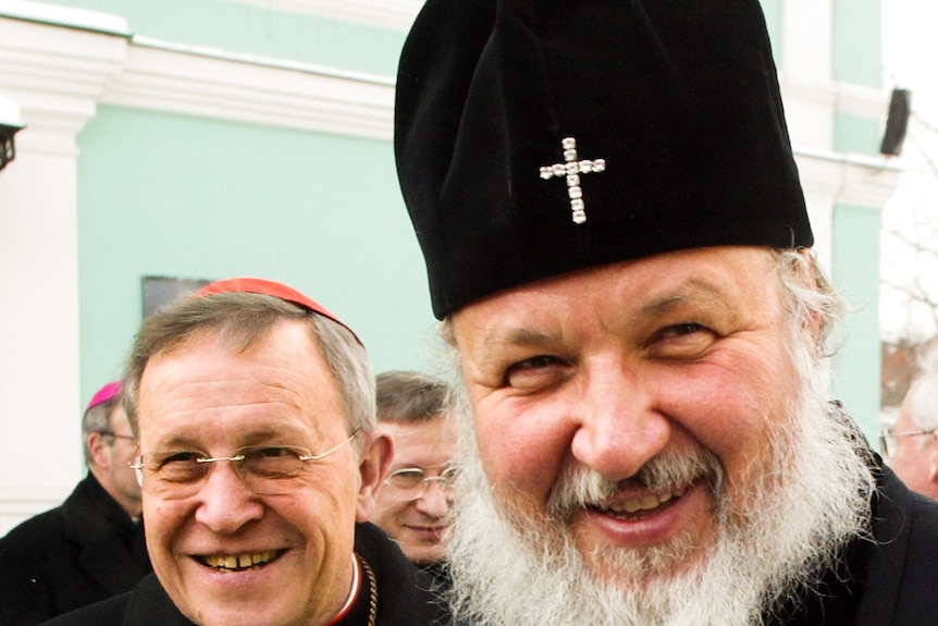 A man wearing a cap and glasses smiles next to Patriarch Kirill dressed in dark robes and a hat with a gold cross.