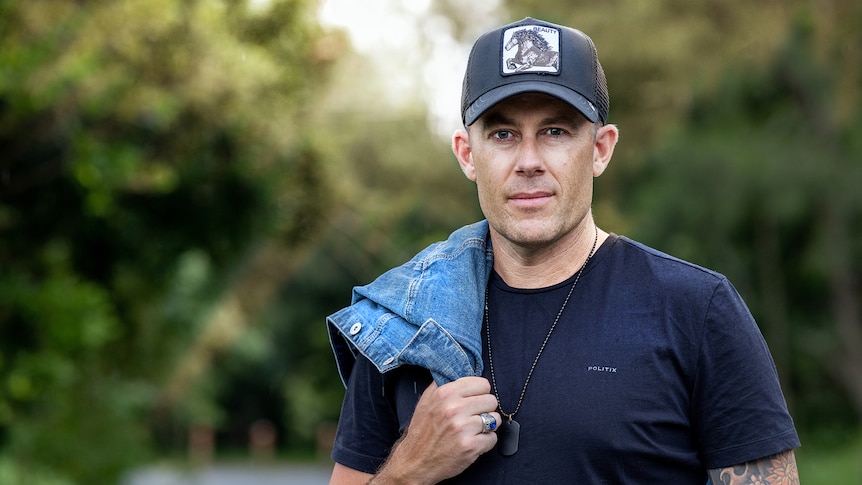 A mid shot of a man wearing a cap in a blue t shirt with a denim jacket draped over his right shoulder and blurred background 