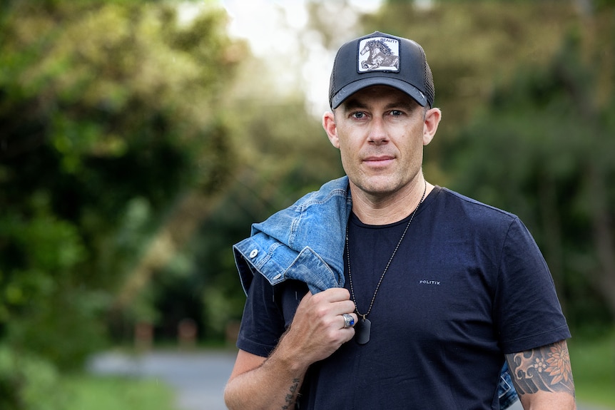 A mid shot of a man wearing a cap in a blue t shirt with a denim jacket draped over his right shoulder and blurred background 