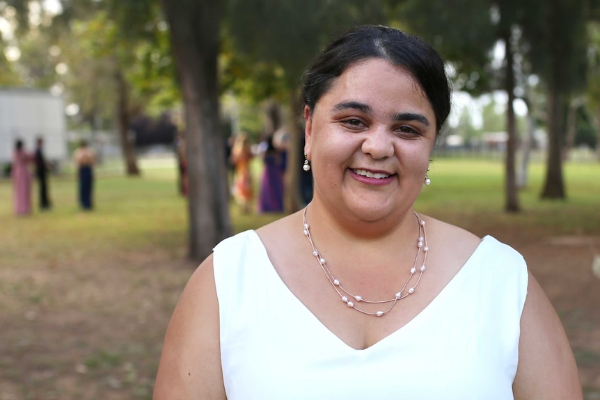 A woman wearing a white dress and a silver jewellery smiles. Behind her are students in formalwear.