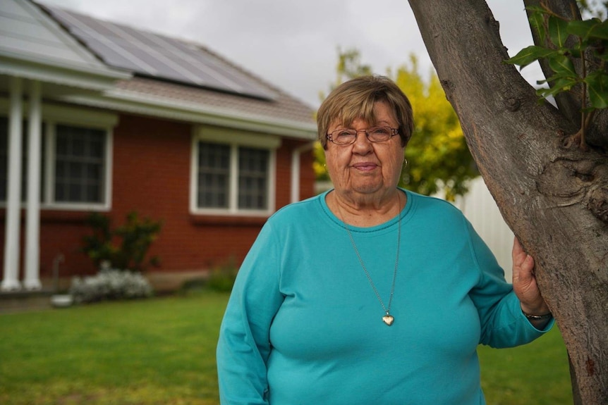 Marianne Hunn, wearing a green-blue top, standing in front of her house, which has solar panels on the roof.