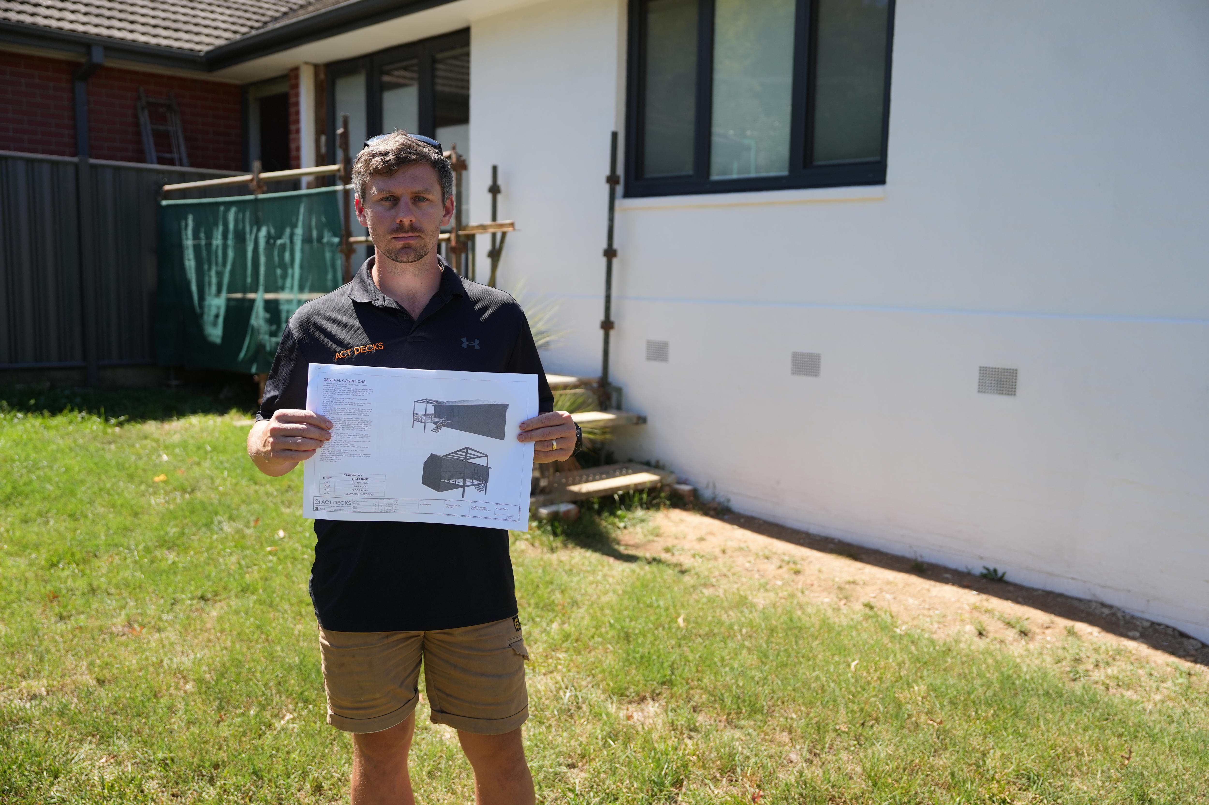 A young man with brown hair stands in front of a white wall with some scaffolding attached, holding plans for a deck