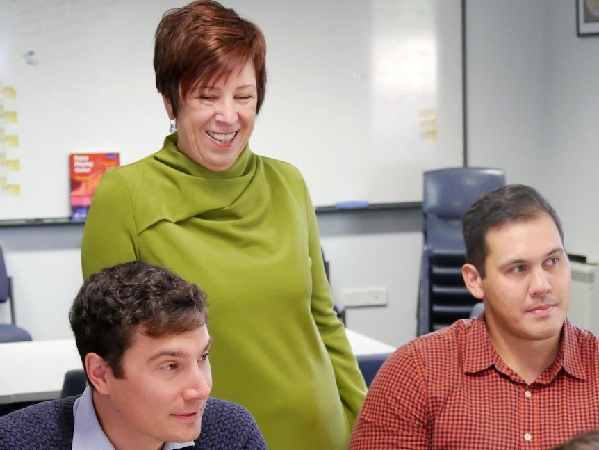 Principal Betty McNeil stands behind two seated men in a classroom.