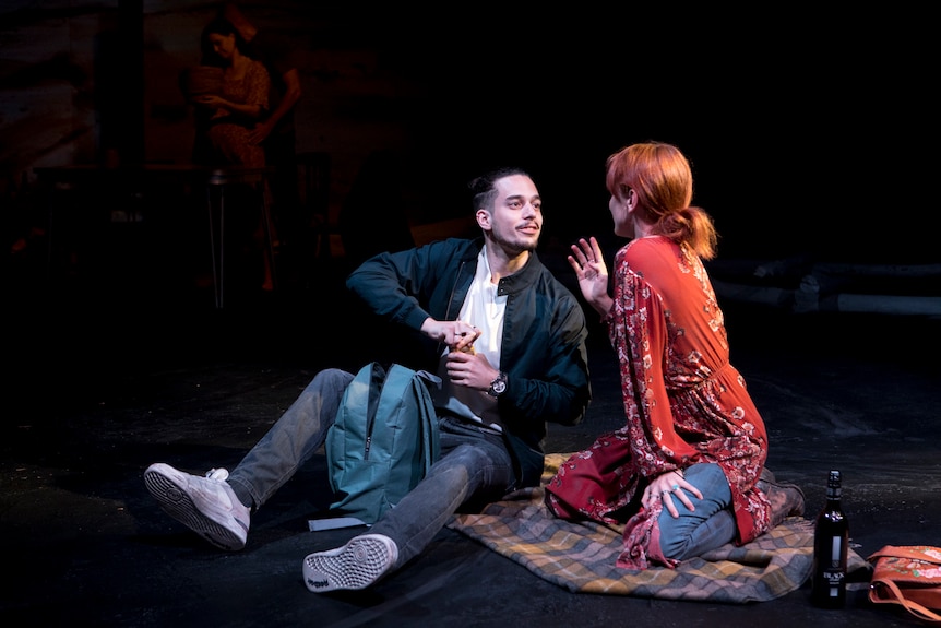 A male and female actor sitting on a blanket on a dark stage. The man is cracking a beer as he listens to the woman talk.