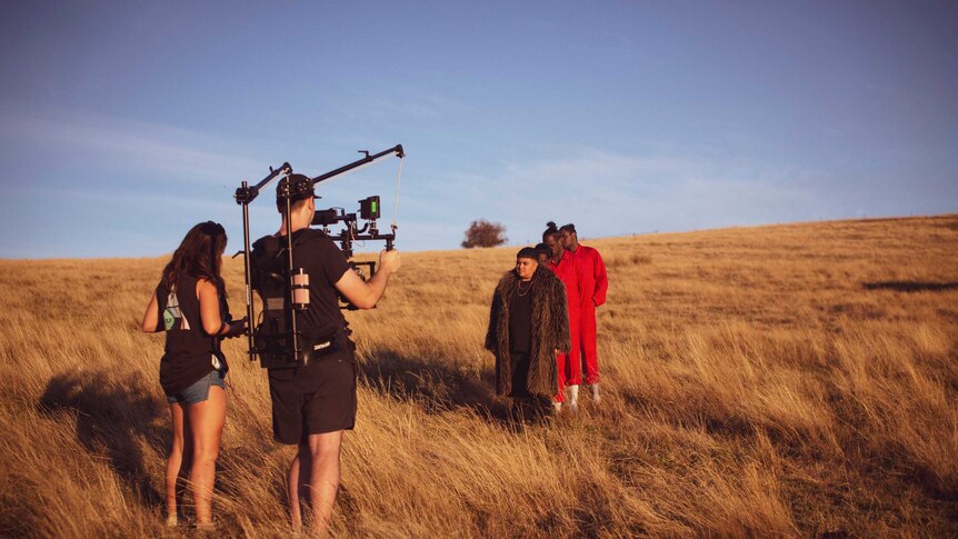 A dry field, a woman stands next to a camera man they direct a group of performers, three in orange jumpsuits
