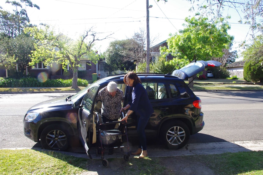 A woman helps her elderly mother out of a black car. Her mother is clutching a walking frame.