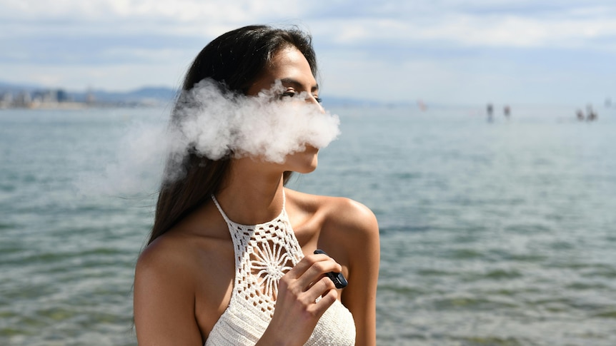 A woman exhaling a cloud of smoke while holding a vape next to the sea.