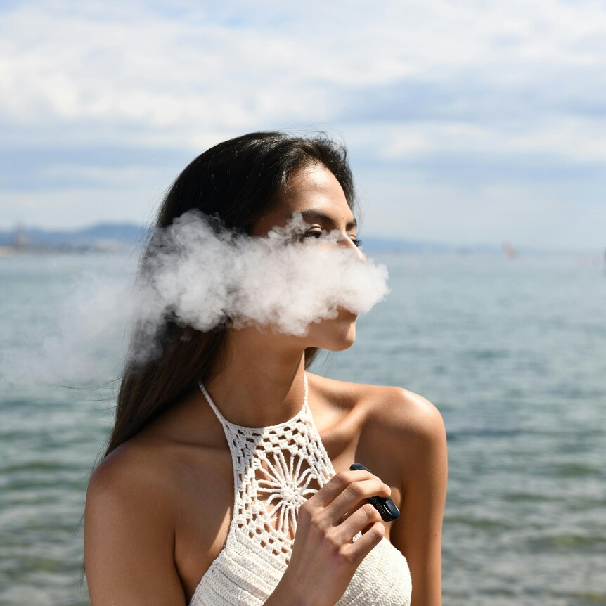 A woman exhaling a cloud of smoke while holding a vape next to the sea.