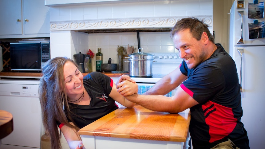 Married couple arm wrestle on kitchen bench.
