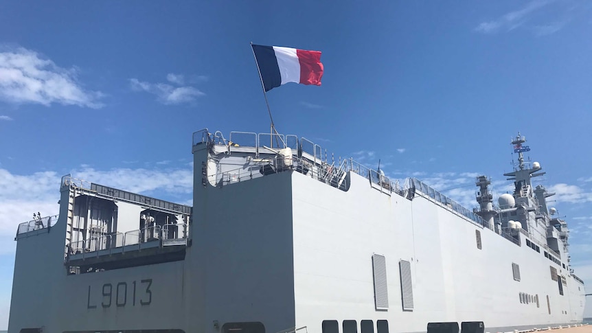 A French warship docked at a harbour on a sunny day with a French flag waving in the wind.