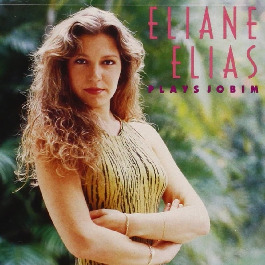 A young Eliane Elias stands in front of some green bushes in a tiger-print, orange dress