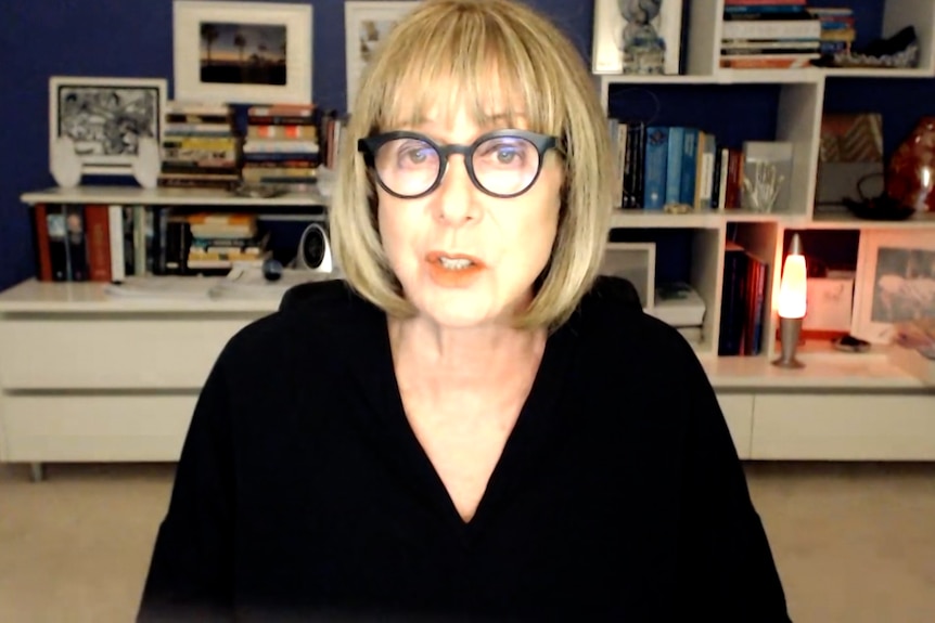 A woman wearing glasses is in a room with books behind her.