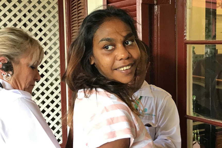 A head and shoulders shot of a young Aborignal woman looking over her shoulder and smiling as guards escort her into court.