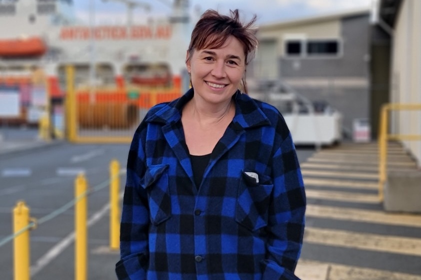 A woman with brown hair stands on a wharf in a blue jacket