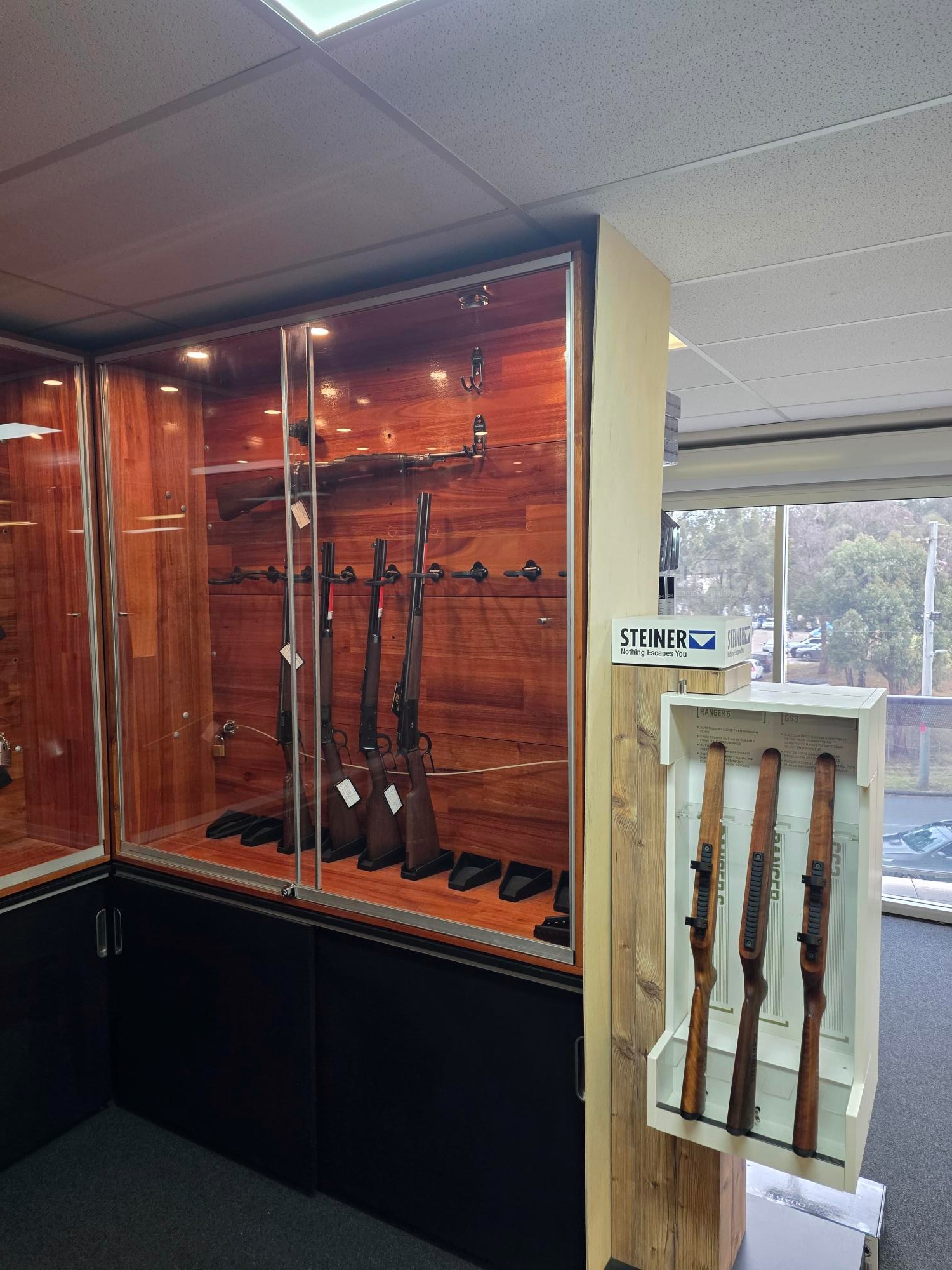 A cabinet with glass dooes with four large rifles standing vertically against the wooden back interior.
