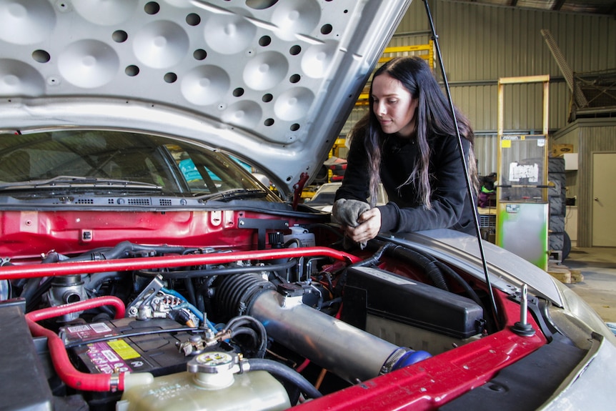 A young woman looks into a car's engine with the bonnet up.