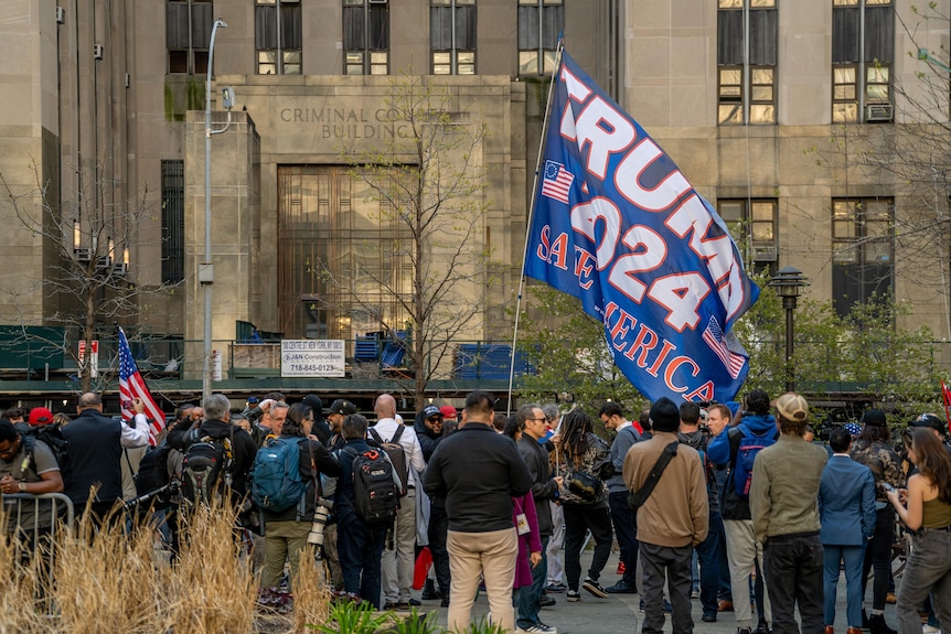 A crowd gathers outside a grey/brown building. Someone holds a large blue "Trump 2024" flag.