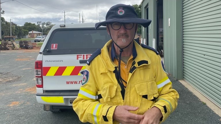 A man in a blue brimmed hat a yellow fire fighters jacket stands in front of an RFS ute.