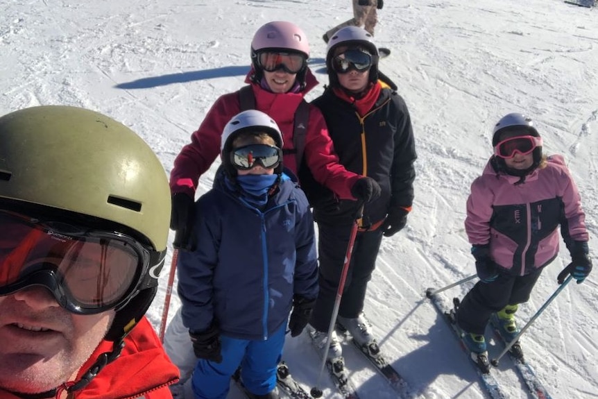 A man wearing skiing clothes and goggles takes a selfie in the snow, his wife and three children stand on skis behind him.