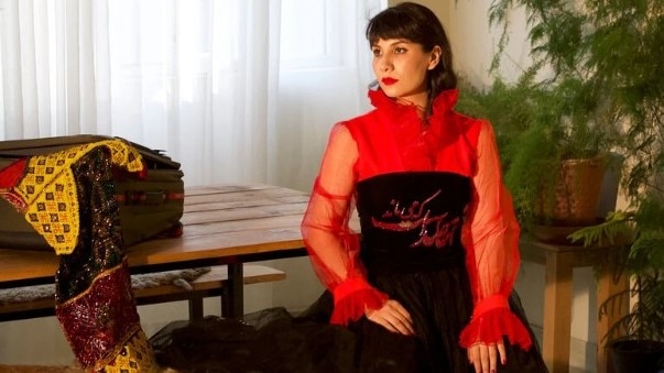 An Afghan woman in red and black dress sits in an apartment filled with plants and beautiful objects