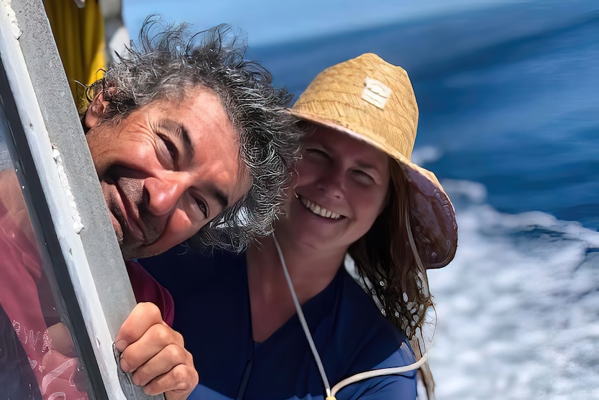A man and woman smile at the camera while travelling on a boat. 