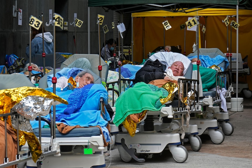 Two elderly patients lie on hospital beds in an outdoor area.