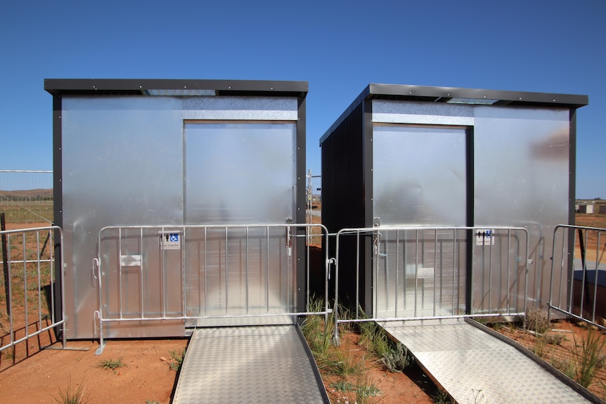 Two metal toilet blocks with gates in front of them on a patch of red dirt with a bare landscape in the background