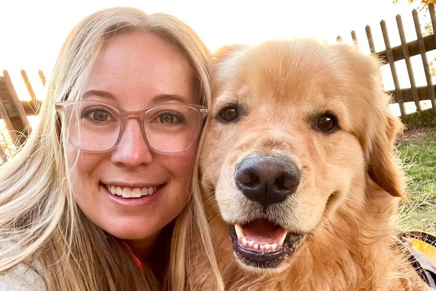 A close up of a blonde woman with glasses smiling with her golden retriever.
