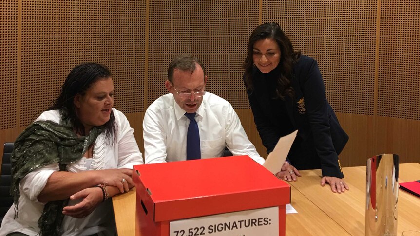 Tony Abbott reads over a piece of paper with two women watching on