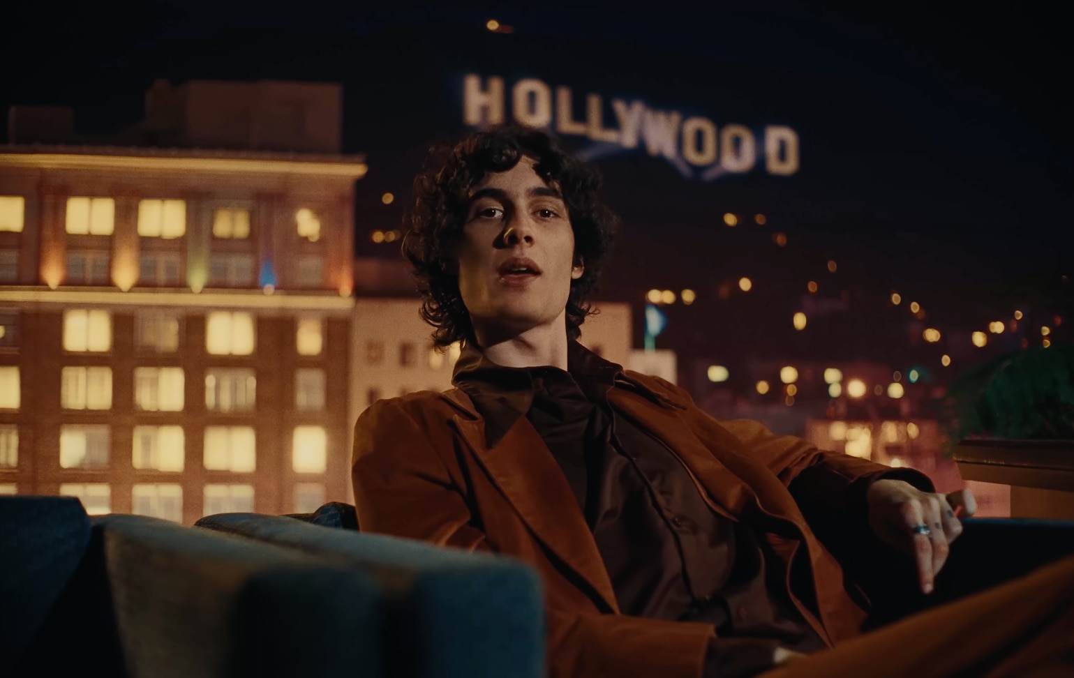 Por la noche, Sombr se sienta en un descapotable con un traje de terciopelo frente al cartel de Hollywood.