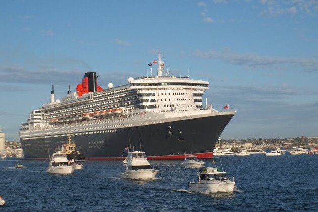 A long shot of the Queen Mary II sailing into harbour with small vessels in front of it.
