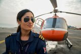 A woman in aviators and pilot outfit stands in front of a red chopper