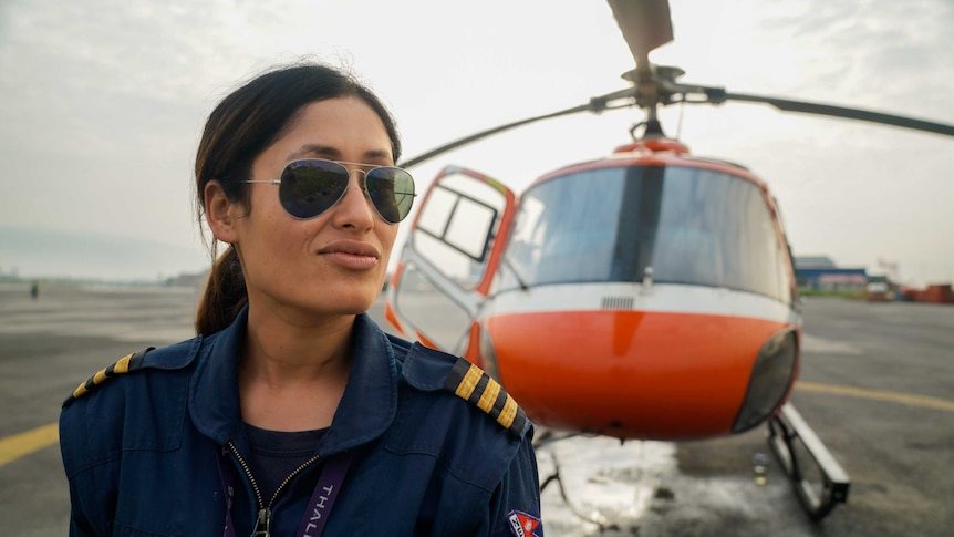 A woman in aviators and pilot outfit stands in front of a red chopper