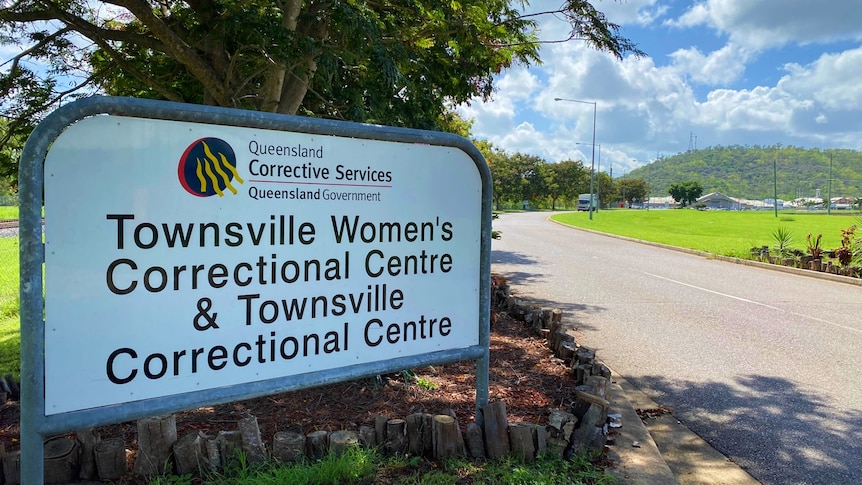 A sign at the entrance to Townsville Correctional Centre in front of a large tree with green grass behind it. 