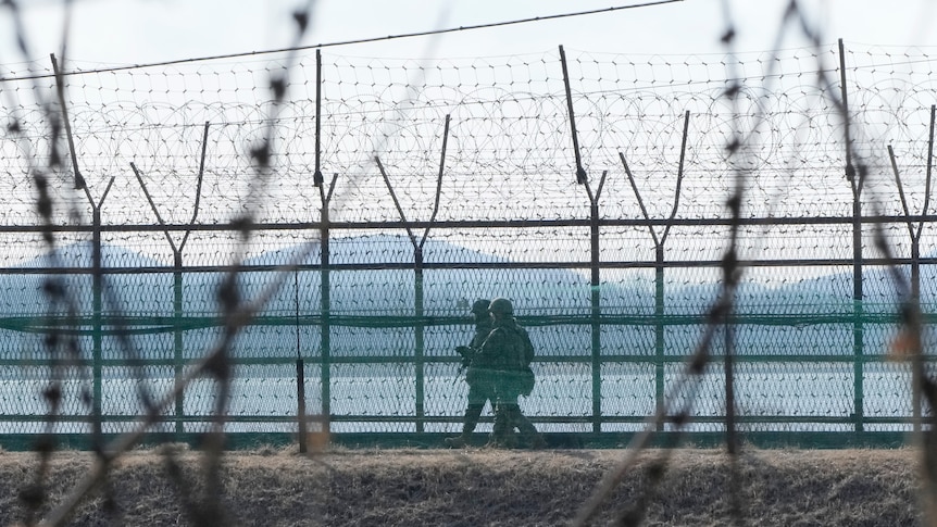 Two armed soldiers in combat gear walk along a path surrounded by many strands of barbed wire, with mountains in the background.
