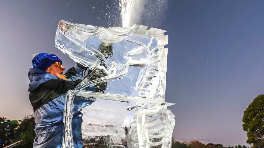 man wearing red overpants and a blue jacket using a power tool to sculpt a block of clear ice that is bigger than him