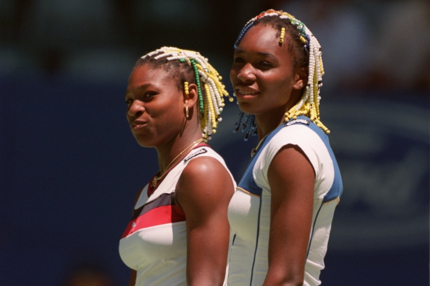 Serena and Venus Williams, with beads in their hair, stand beside each other on a tennis court.