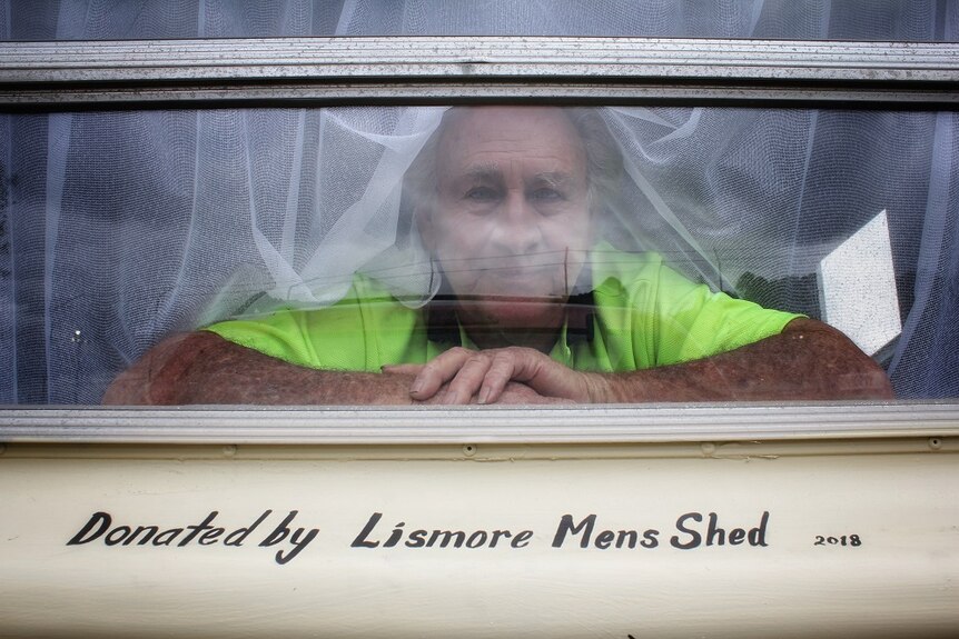A man peers through the back window of a caravan with the words 'donated by the Lismore Mens Shed' below.