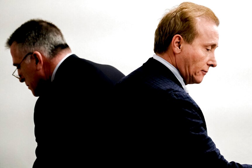 Two men wearing suits sit back-to-back.