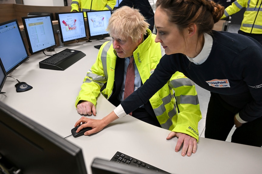 Boris Johnson an einem Schreibtisch mit Computerbildschirmen in einem Kernkraftwerk, während ein Mitarbeiter mit einer Maus auf seinem Bildschirm navigiert