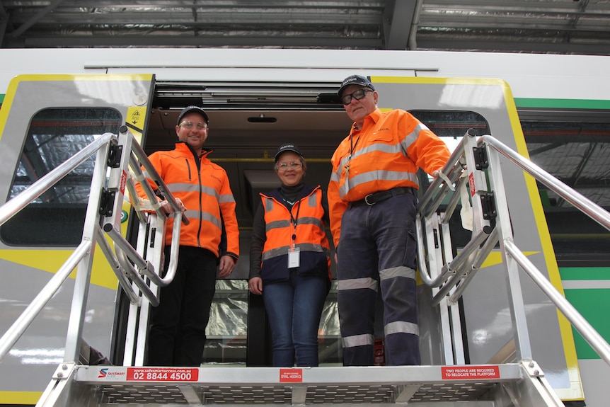Three workers pictured from below wearing high-vis and standing in the doorway of a new Metronet train