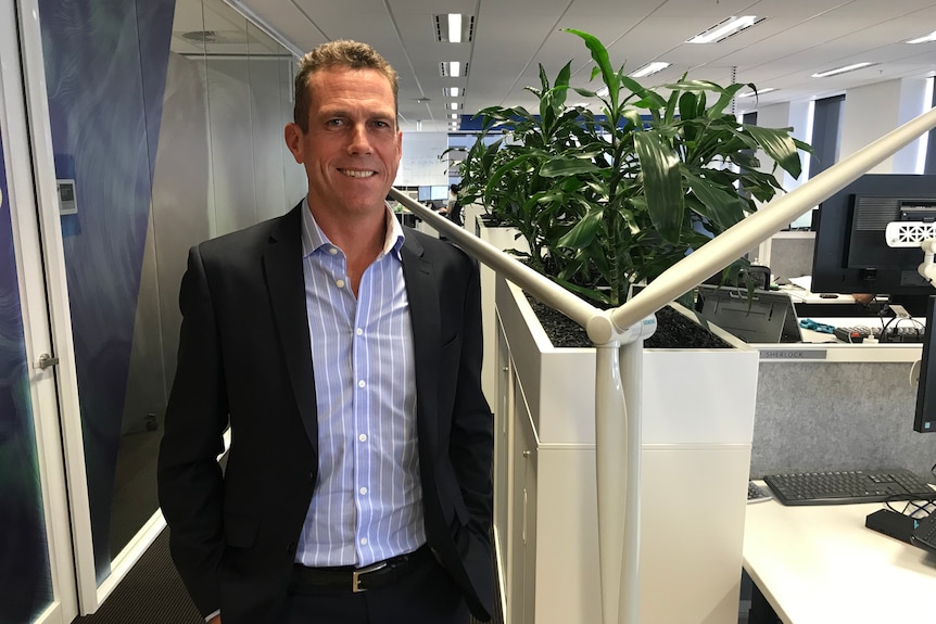 A man with a suit jacket over a blue and white striped business shirt stands in an office near a human-sized wind turbine.