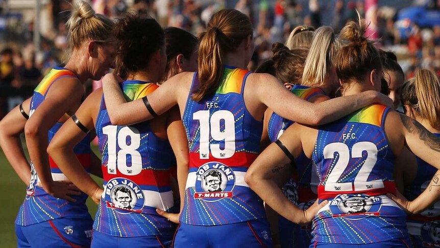 A group of women in sports jerseys with rainbow on them stand in a huddle