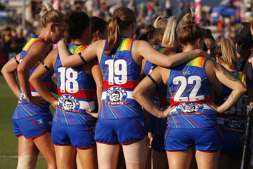 A group of women in sports jerseys with rainbow on them stand in a huddle