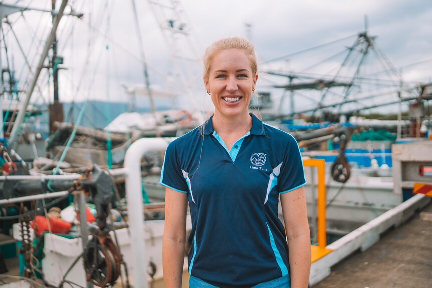 A smiling blonde woman stands in front of tuna boats.