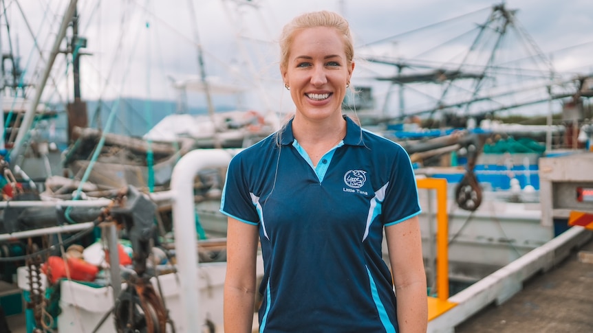 A smiling blonde woman stands in front of tuna boats.
