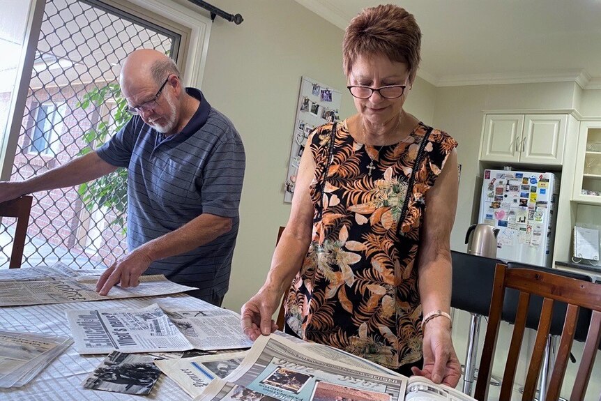 Two people reading newspaper clippings on a kitchen table. They are both looking down.