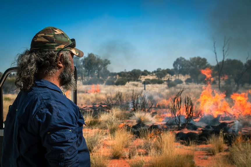 A man wearing a cap looks out at a dessert landscape which is being burnt by orange flames