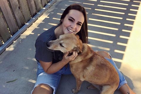 Woman with ripped jeans and grey top sits on ground with a dog